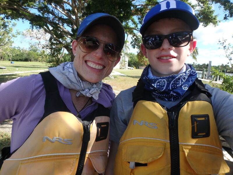 The image shows a woman and a child, likely a mother and son, posing for a selfie outdoors. Both are wearing life vests, suggesting they are about to engage in a water activity like kayaking or boating. The woman is on the left, wearing a blue cap and sunglasses, while the child is on the right, also wearing a blue cap and sunglasses. They are both smiling at the camera. The background features trees and a glimpse of water, indicating a natural setting.